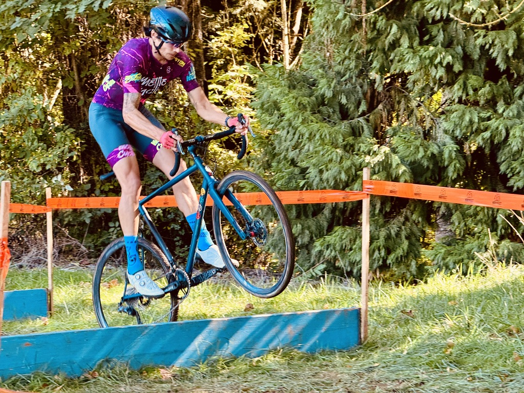 A rider in purple and teal lifts their bike over a blue cyclocross barrier.