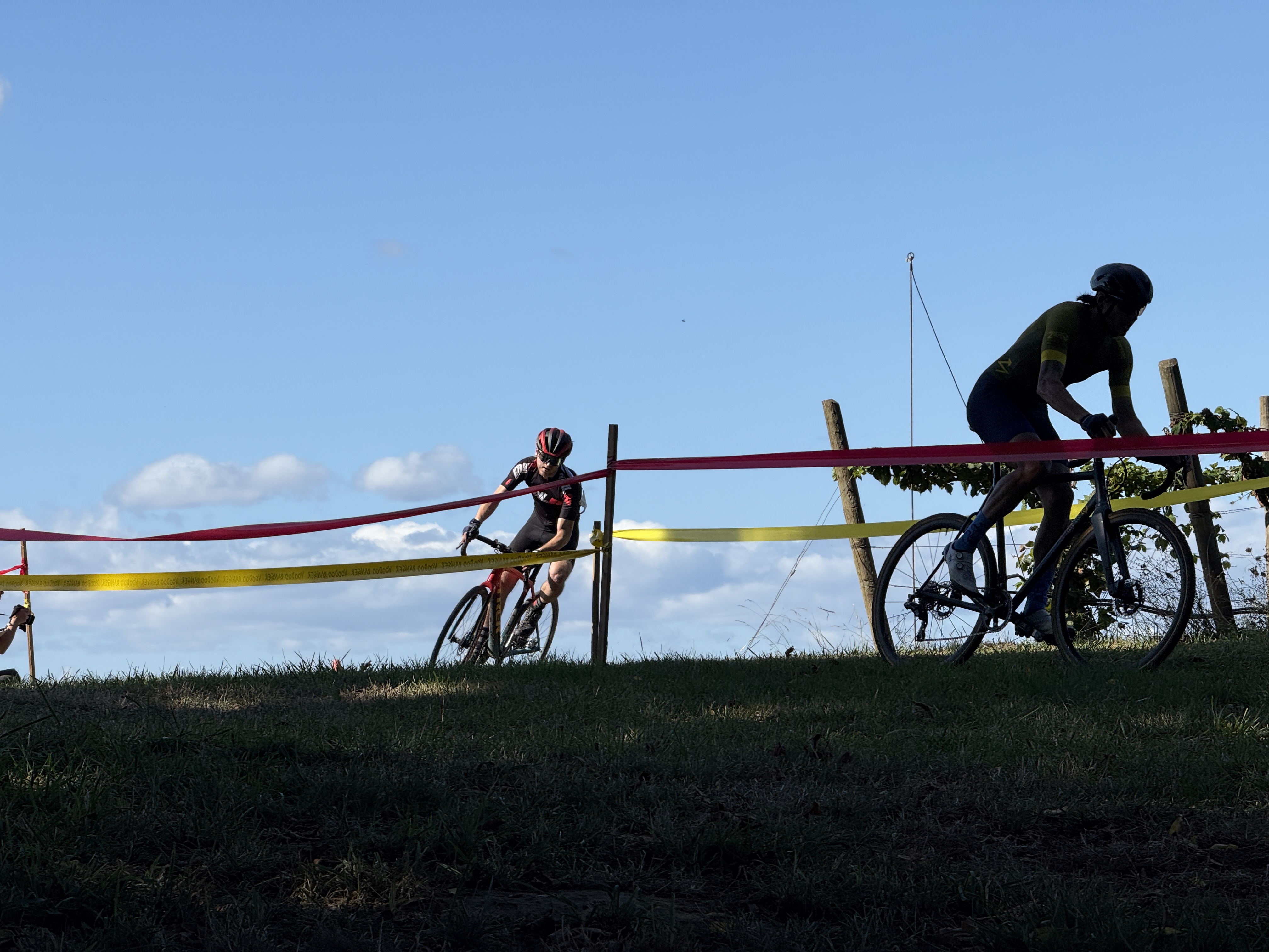 Two cyclocross riders crest a grassy hill between red and yellow course tape under a blue sky.