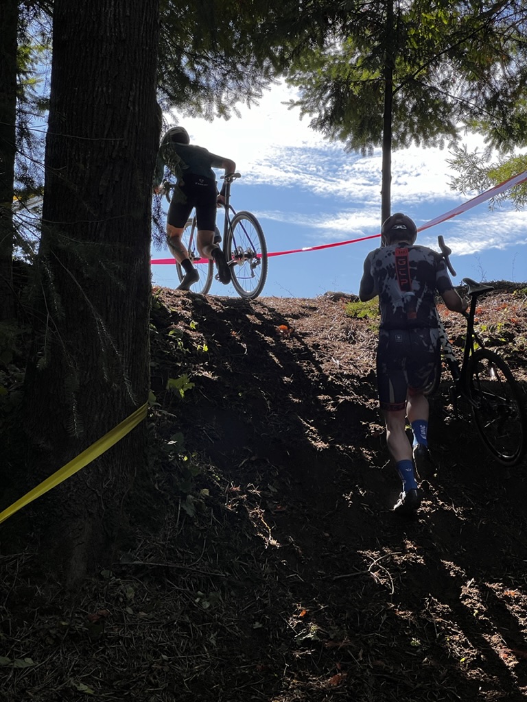 Two racers carry their bikes up a steep, wooded run-up.